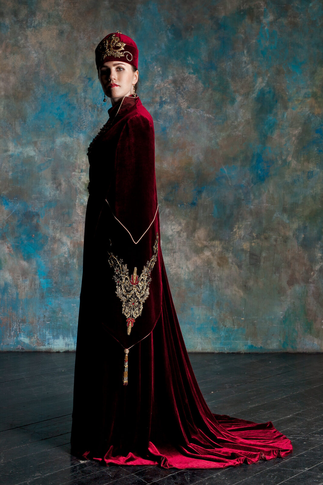 A full-length portrait of Nafset in traditional Circassian dress — a deep red velvet coat-dress with a long, dramatic train pooling on the floor, gold embroidery running down the front and along the hem, and a matching red traditional hat crowned with gold ornamentation. She stands in three-quarter profile against a softly textured blue-grey backdrop.