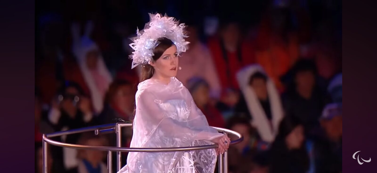 A screen capture from the XI Winter Paralympics Closing Ceremony broadcast. Nafset stands on a raised, illuminated round platform encircled by a low metal railing, both hands resting on the rail. She wears an elaborate white performance costume — a sheer, iridescent wrap over a voluminous, structurally pleated white gown — with a crown-like headpiece of white feather-like spikes and small lights woven through her hair, and a headset microphone at her cheek. Behind her, a packed stadium audience bundled in winter clothing, many in red and orange, is visible under the ceremony’s dim, colorful lighting.