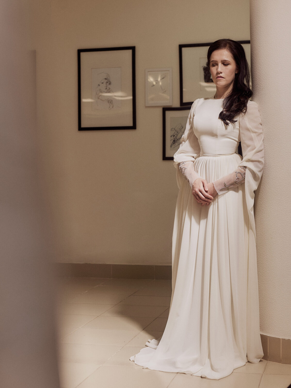 Nafset stands leaning gently back against a wall, wearing a flowing ivory gown with sheer, embroidered sleeves visible at the wrists. Her hands are clasped in front of her, her expression calm and contemplative, softly lit in three-quarter shadow.