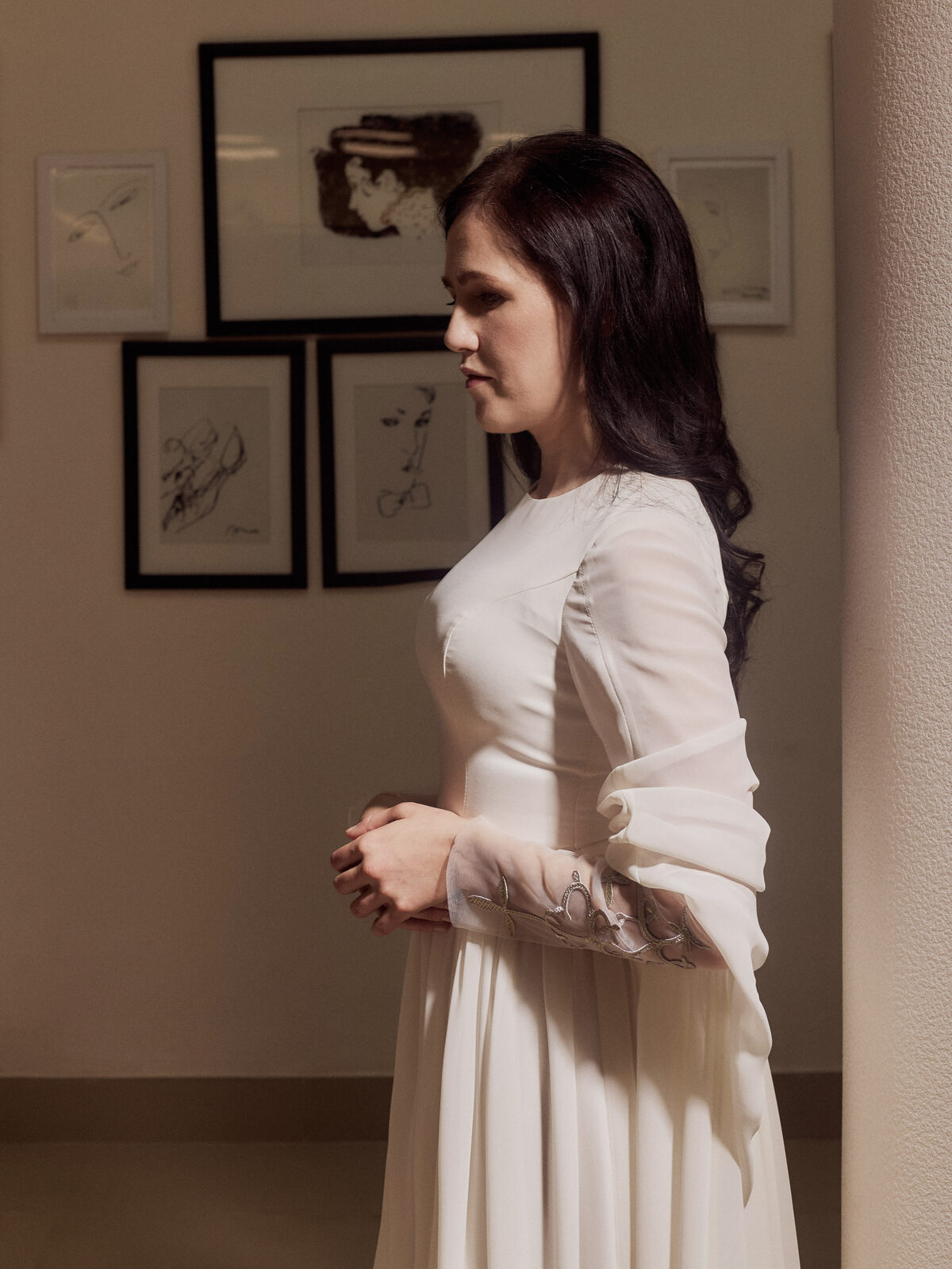 Nafset stands in a three-quarter turn, wearing a flowing ivory floor-length gown with long sleeves, one hand resting lightly at her waist. Her dark wavy hair falls loose over one shoulder. She stands in a softly lit room with black-framed line-art sketches on the wall behind her.