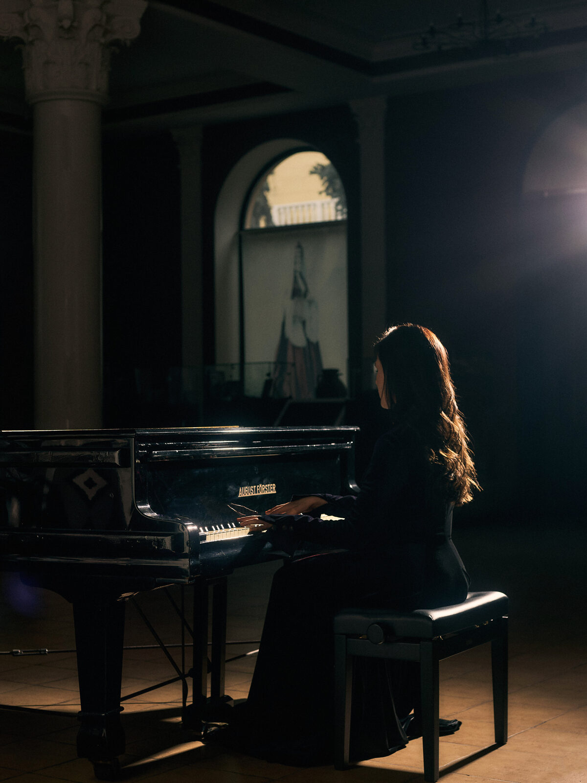 A striking silhouette-style portrait of Nafset in profile, seated at a piano and wearing a dark, high-necked outfit, her hair falling in loose waves. Dramatic side lighting traces the line of her face and shoulder against a darkened background, evoking the atmosphere of a live performance.