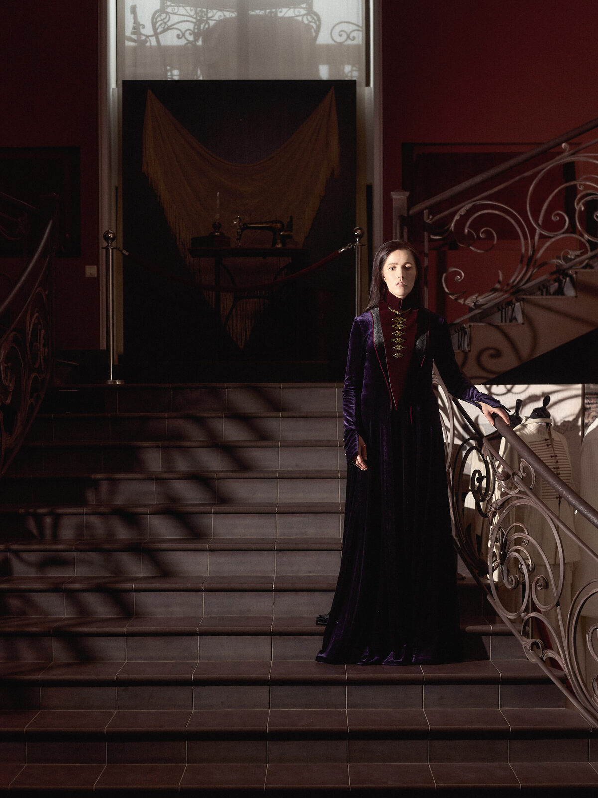 Nafset stands partway up an ornate stone staircase with wrought-iron railings, one hand resting on the banister. She wears a deep purple velvet gown with an ornate embroidered collar in burgundy and black, her expression composed and poised, in a grand, dimly lit interior.