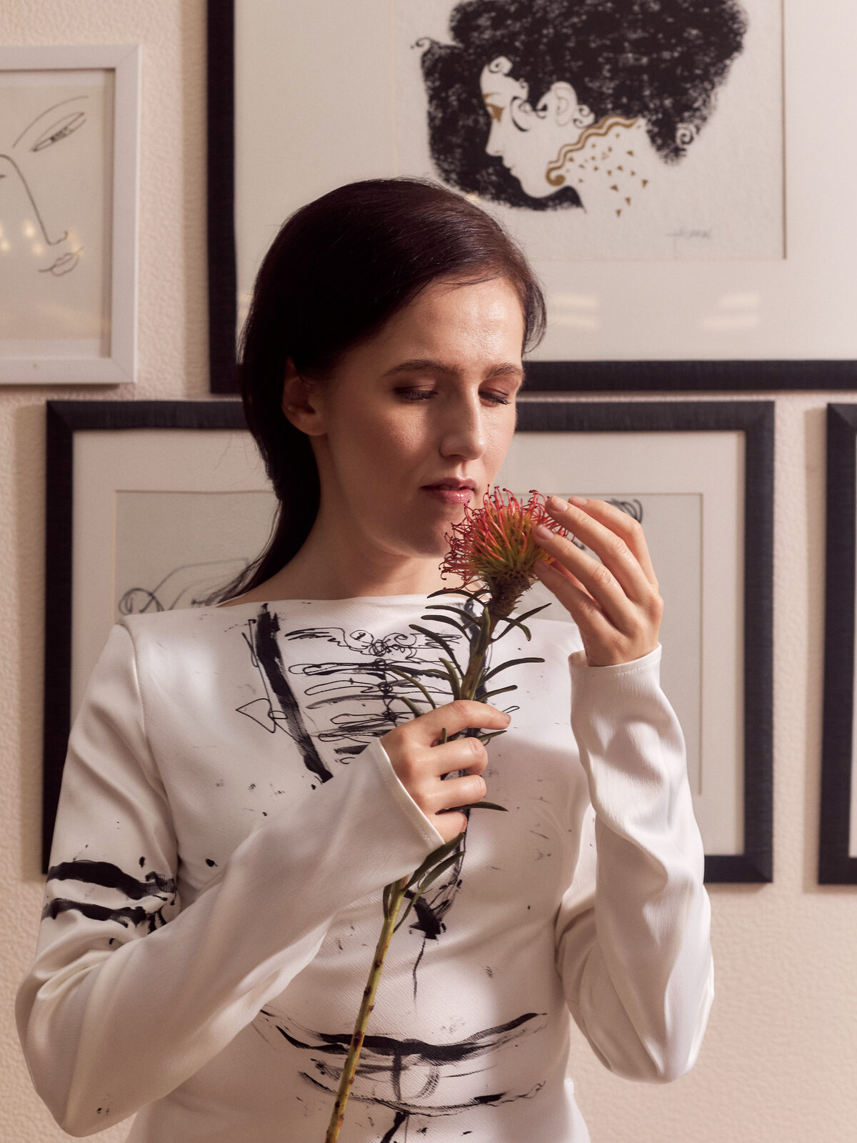 A close, intimate portrait of Nafset holding a single reddish flower near her face, her eyes open and gaze soft. She wears a white dress with a bold, abstract black brushstroke design across the bodice and sleeve, standing before a wall of framed line drawings.